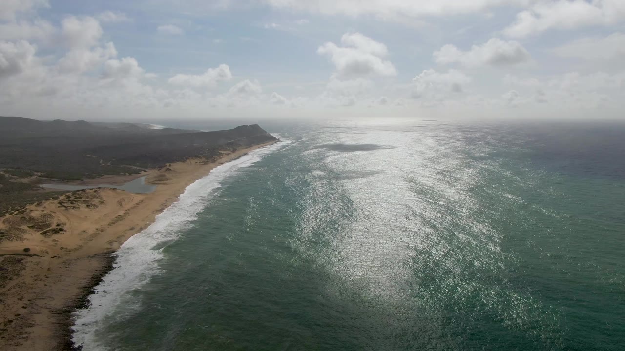 At 0:01, the Punta Gorda East Cape shoreline remains in a high aerial view as white foam shifts along the beach beside dry hills and no visible people.