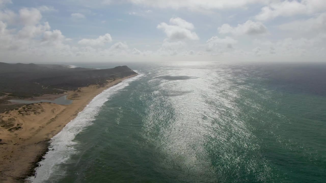 Aerial coastline view with white waves, sand, hills, and clouds.