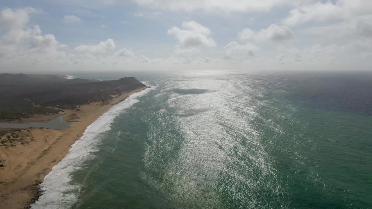 Aerial view at Punta Gorda, East Cape, Baja California Sur with foamy waves advancing toward the sand, strong reflected light across the ocean surface, the inlet still visible at left, and no visible people.