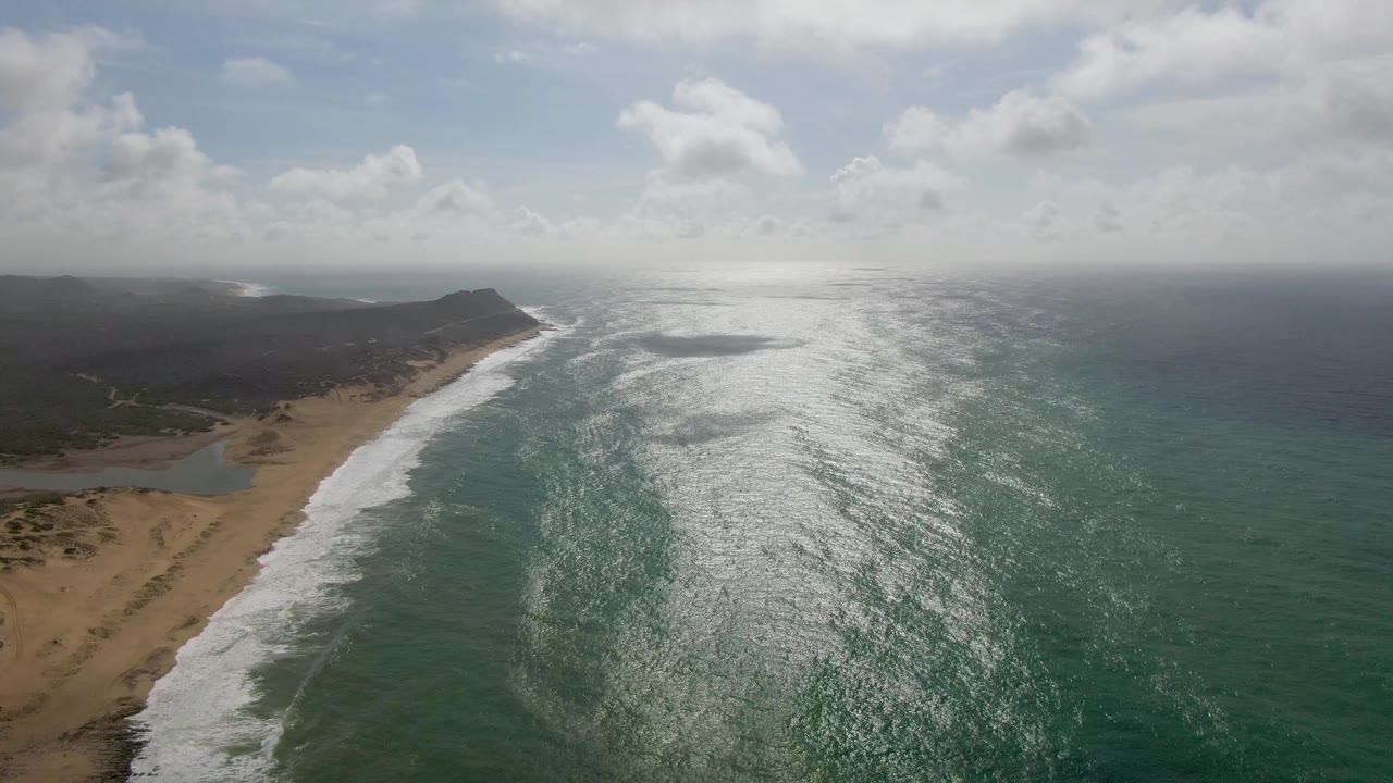 Drone-style frame over Punta Gorda, East Cape, Baja California Sur showing the surf band slightly farther along the curved sand, open ocean filling the right side, inland terrain in shadow, and no visible people.