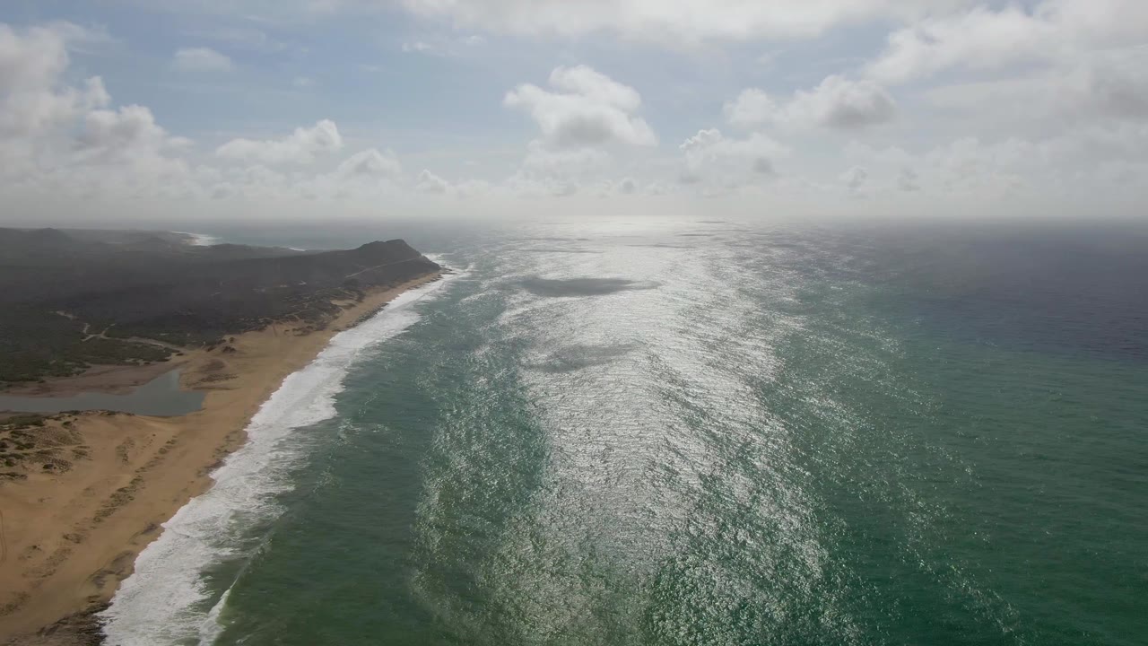 Aerial frame from Punta Gorda, East Cape, Baja California Sur with the sandy beach and inlet more prominent at left, white breakers moving shoreward, bright reflected light across the water, and no visible people.