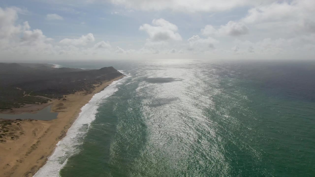 Aerial view of waves breaking along a sandy coastline under scattered clouds.
