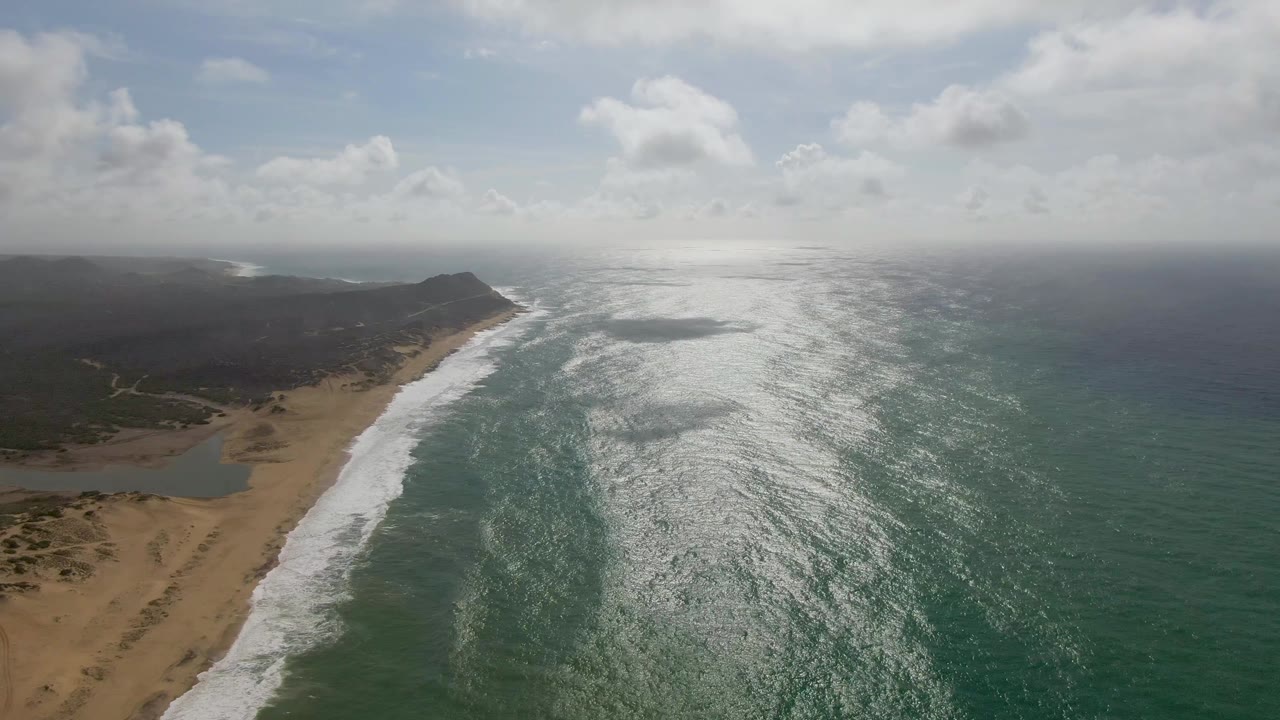 Drone frame from Punta Gorda East Cape showing a diagonal sandy shoreline, advancing white foam, a small inland water area near the lower left, bright ocean surface, and no visible people.