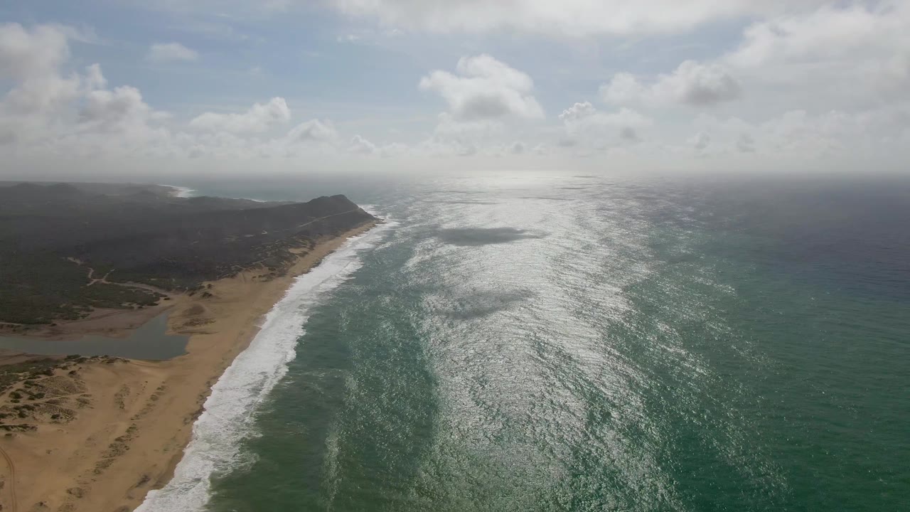 Wide aerial frame at Punta Gorda East Cape with foamy waves along the beach, darker land stretching toward a low headland, cloud shadows over the coast, bright water to the right, and no visible people.