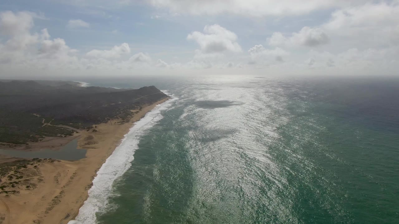 Aerial view over Punta Gorda East Cape with a broad strip of white surf, sandy dunes at lower left, a small lagoon-like water patch behind the beach, open ocean to the right, and no visible people.