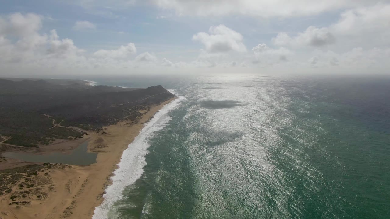 Aerial frame from Punta Gordo, Baja California Sur showing white surf running along a sandy beach, darker inland vegetation, bright ocean glare, and no visible people.