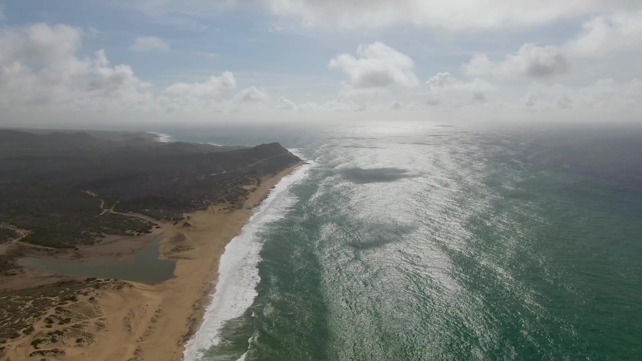 Aerial view over Punta Gordo with foamy waves tracing the beach, open ocean filling the right side, low inland terrain to the left, and no visible people.