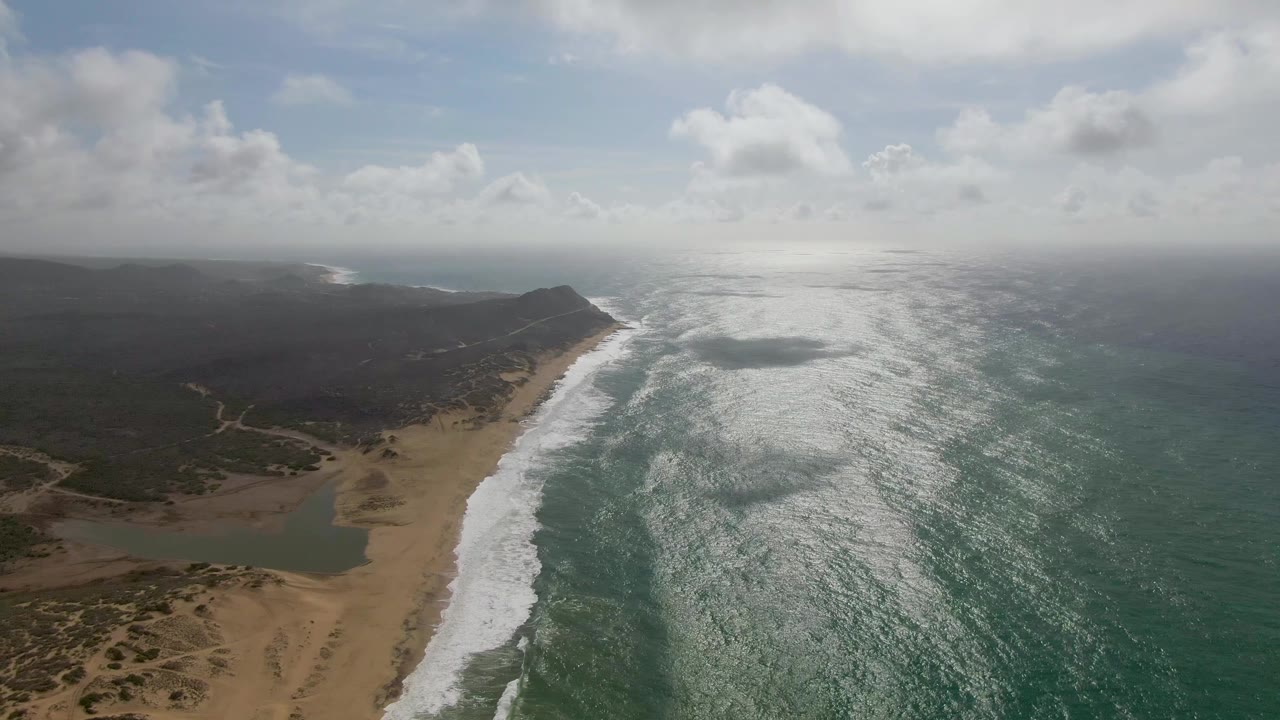 Wide aerial beach view with surf, ocean glare, and dark coastal vegetation inland.