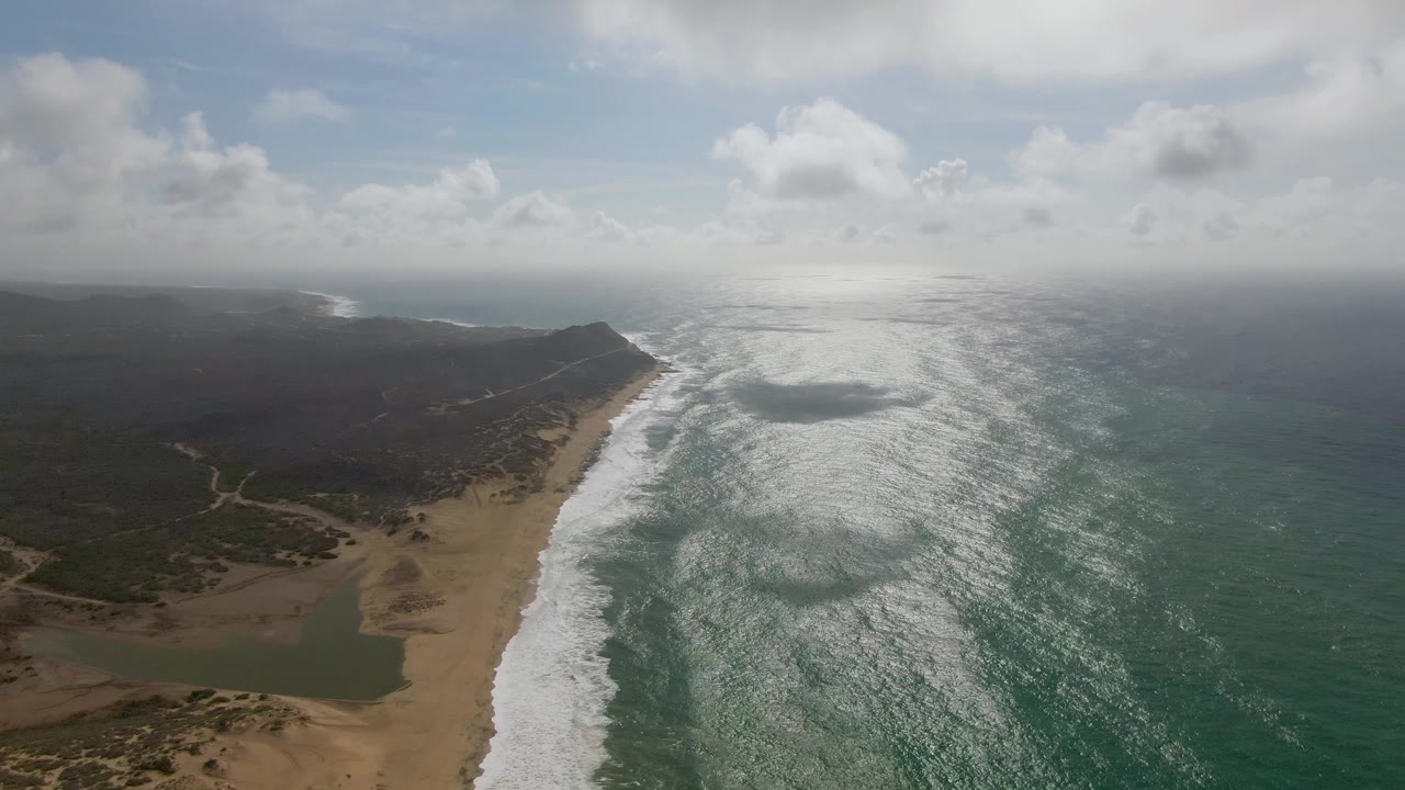 A wide Punta Gorda coastal view shows the surf line curving past the beach toward dark low hills under broken clouds, with no visible people on the sand or water.