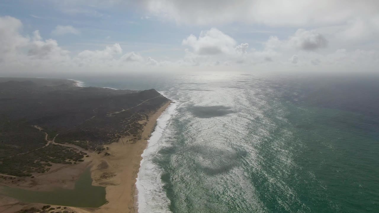 At 0:00, an aerial view over Punta Gorda East Cape shows white surf meeting a sandy beach, green-blue sea to the right, dark vegetation and low hills to the left, and no visible people.