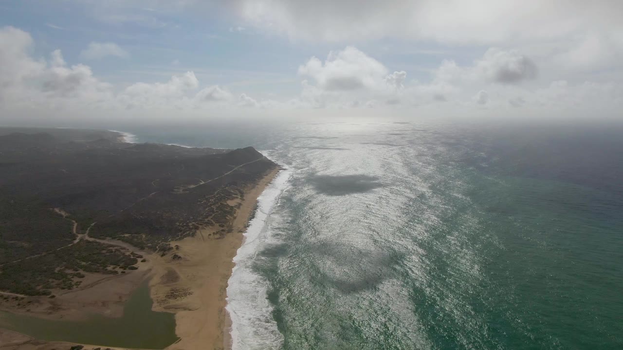 At 0:01, foamy wave lines push farther along the sandy Punta Gorda East Cape shore while the ocean surface brightens under hazy cloud light, with no visible people.