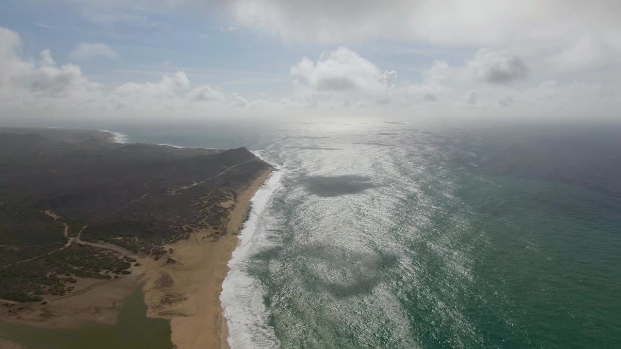 At 0:02, the drone view keeps the surf line centered between the tan beach and green ocean, with dark inland terrain and low hills receding into haze and no visible people.