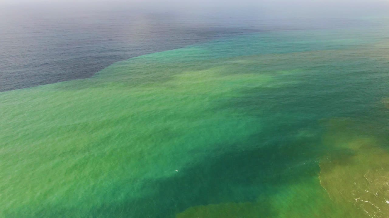 Aerial view at Punta Gorda East Cape where clear turquoise water reveals submerged sand tones and a darker patch near the center, with no visible people.