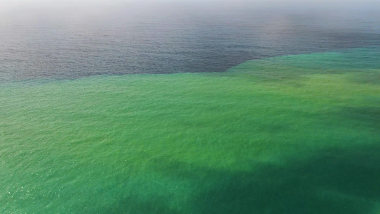 Aerial view over Punta Gorda shallow turquoise-green water blending into darker blue-gray ocean near the hazy horizon, with no visible people.