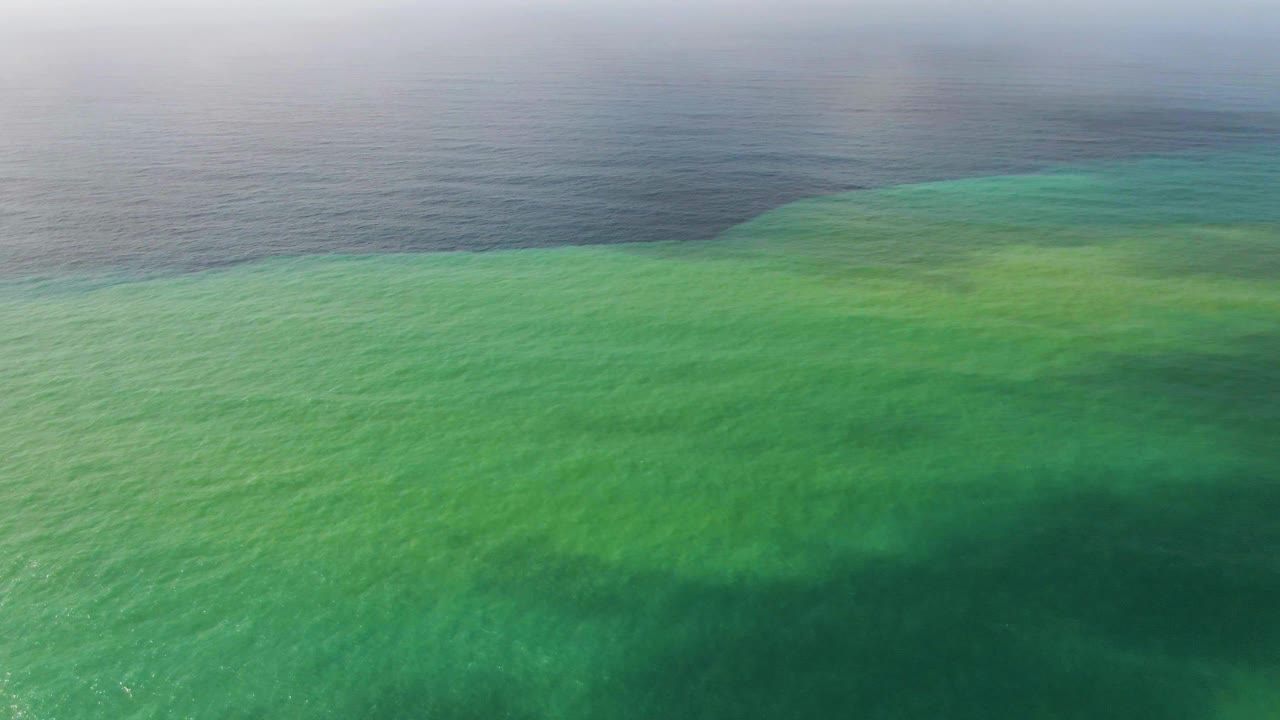 Aerial coastal water at Punta Gorda shows shallow green texture fading into blue-gray open ocean under diffuse haze, with no visible people.