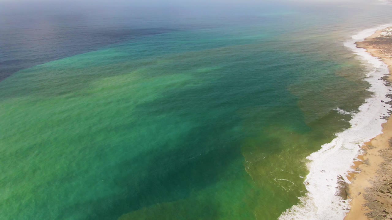 Punta Gorda coastal aerial showing turquoise water across most of the frame, white waves along the shore, and more exposed sandy terrain at the far right, with no visible people.