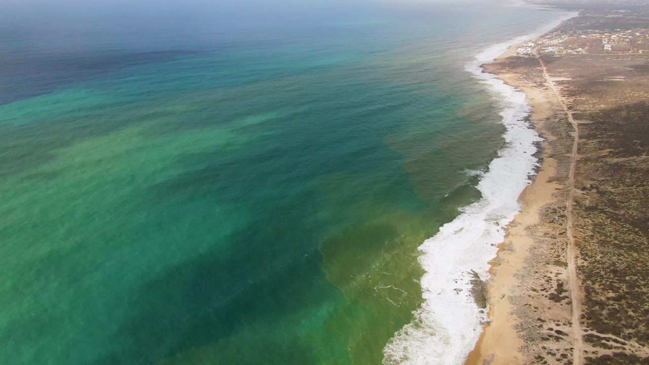 Aerial view over Punta Gorda, Baja California Sur, with turquoise ocean on the left, white surf along the sandy shore, arid scrubland on the right, a parallel dirt road, and no visible people.