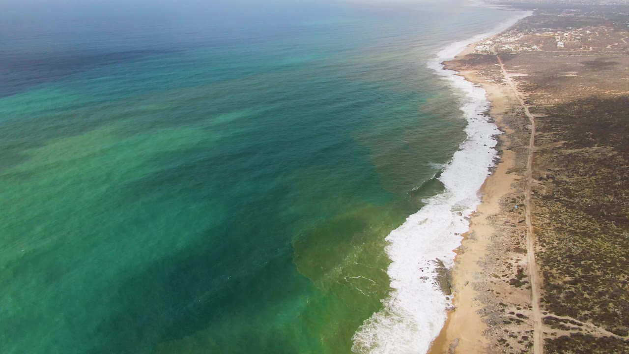 Aerial frame over Punta Gorda showing green-blue water and a darker shadow patch offshore, white waves breaking onto sand, dry coastal terrain, a dirt road, and no visible people.