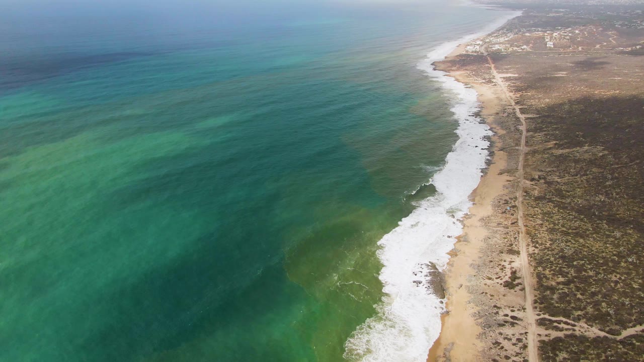 High aerial view of Punta Gorda coastline with a broad turquoise water field, a bright white surf band along the beach, scrubland to the right, a coastal dirt track, and no visible people.