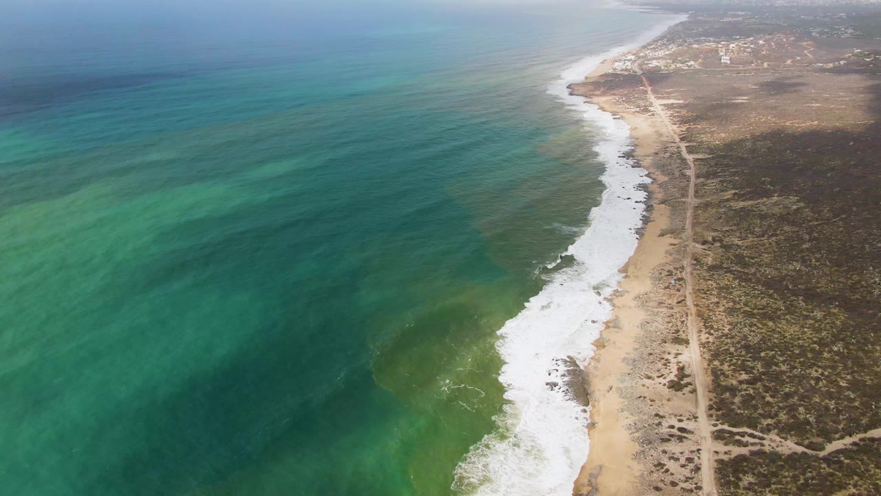 Aerial coastal scene with ocean shadow, white surf, sand, scrubland, and road.