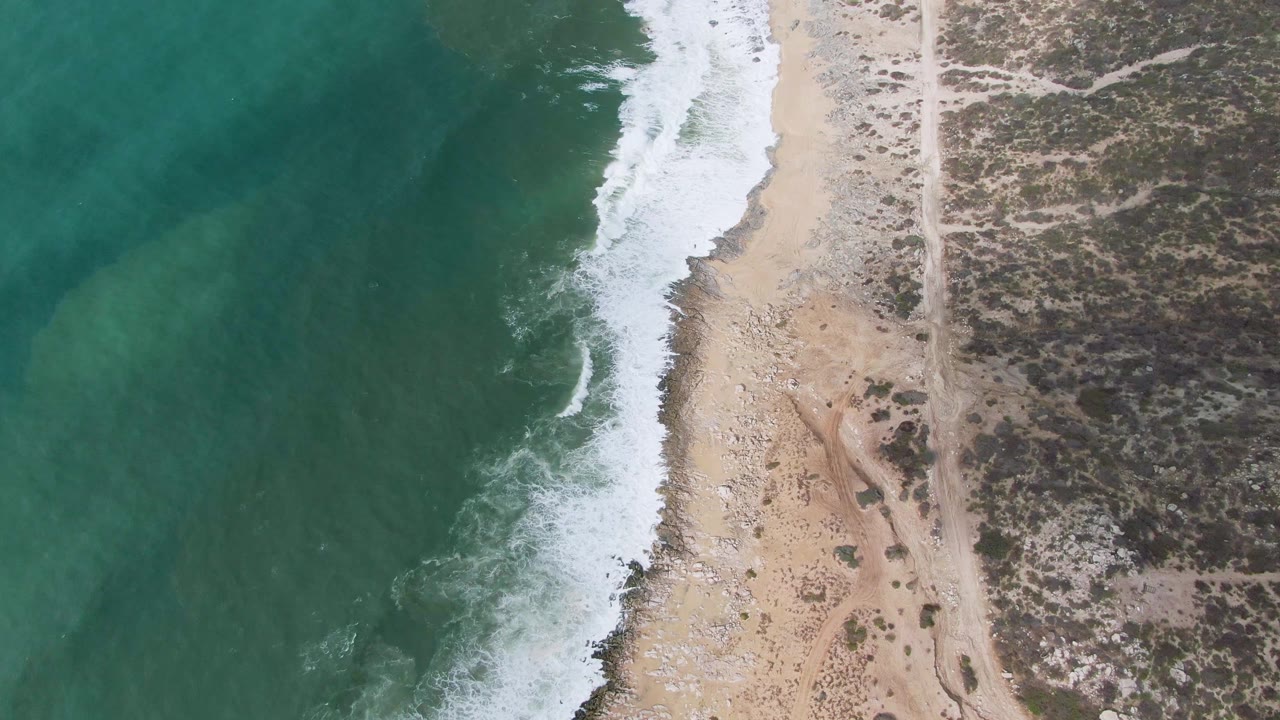 Overhead shoreline with turquoise water, white surf, sandy beach, dirt road, and scrubland.