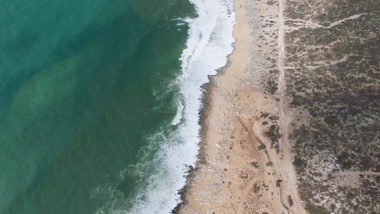 Aerial frame from Punta Gordo, Baja California Sur, showing the white wave band shifted along the beach, green-blue water offshore, pale sand and pebbles at center, scrubland inland, and no visible people.