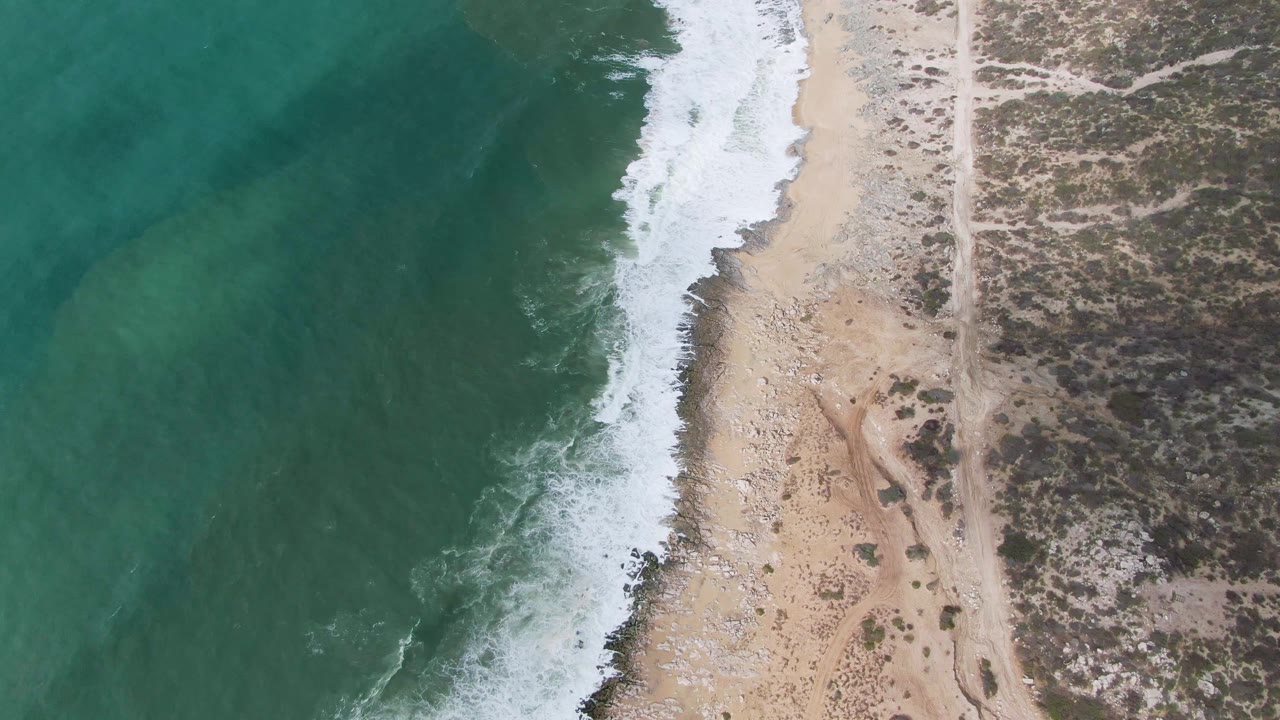 Overhead view of the Punta Gordo shoreline with a broader white foam strip along the sand, darker turquoise water offshore, a narrow inland dirt road, dry scrubland, and no visible people.