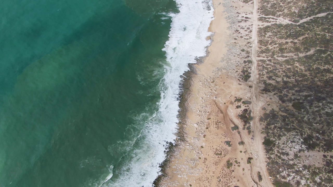 Mid-clip aerial frame over Punta Gordo with green water pushing a fresh line of foam onto the sandy beach, scattered pebbles near the shore, a dirt road inland, scrubland at right, and no visible people.