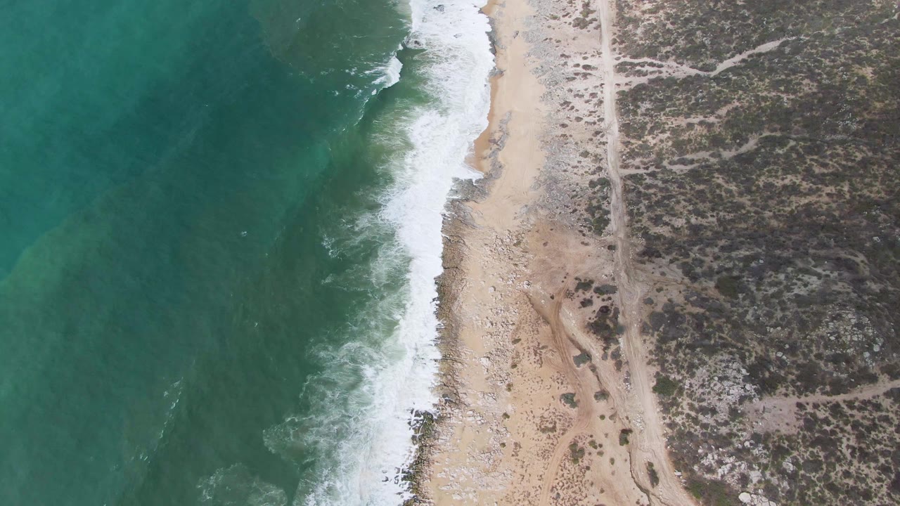 Overhead view at Punta Gorda, East Cape, Baja California Sur, with turquoise water, white surf, pale sand, a dirt path, scrubland, and no visible people.