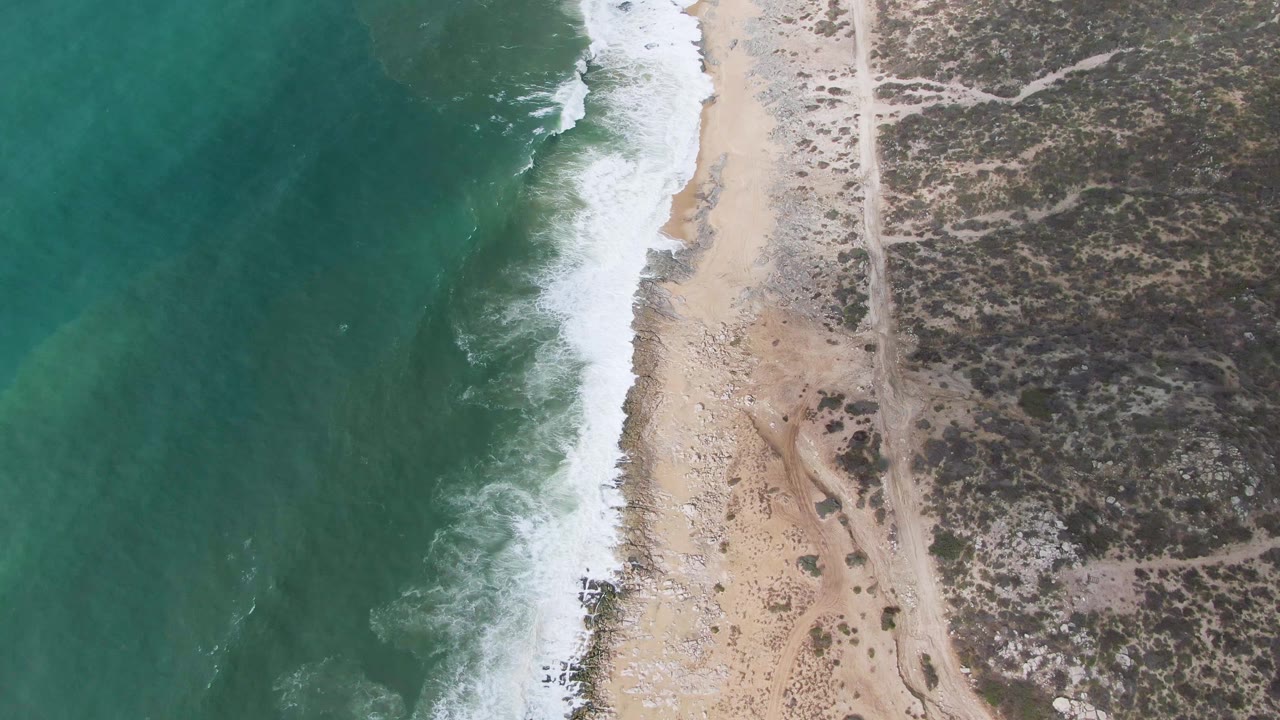 A later overhead moment at Punta Gorda shows white foam pushing along the sand, green water to the left, a parallel dirt path to the right, scrub vegetation, and no visible people.