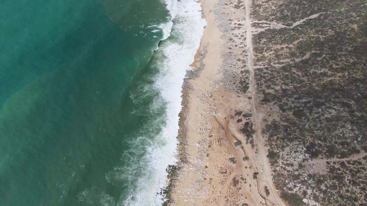 Overhead Punta Gorda coastline frame with darker green water on the left, a bright foamy break near the sand, sparse beach rocks, inland scrub, and no visible people.