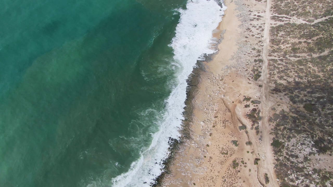 Aerial frame over Punta Gorda, Baja California Sur showing turquoise water on the left, white surf along a sandy and rocky shoreline, a dirt coastal road on the right, and no visible people.