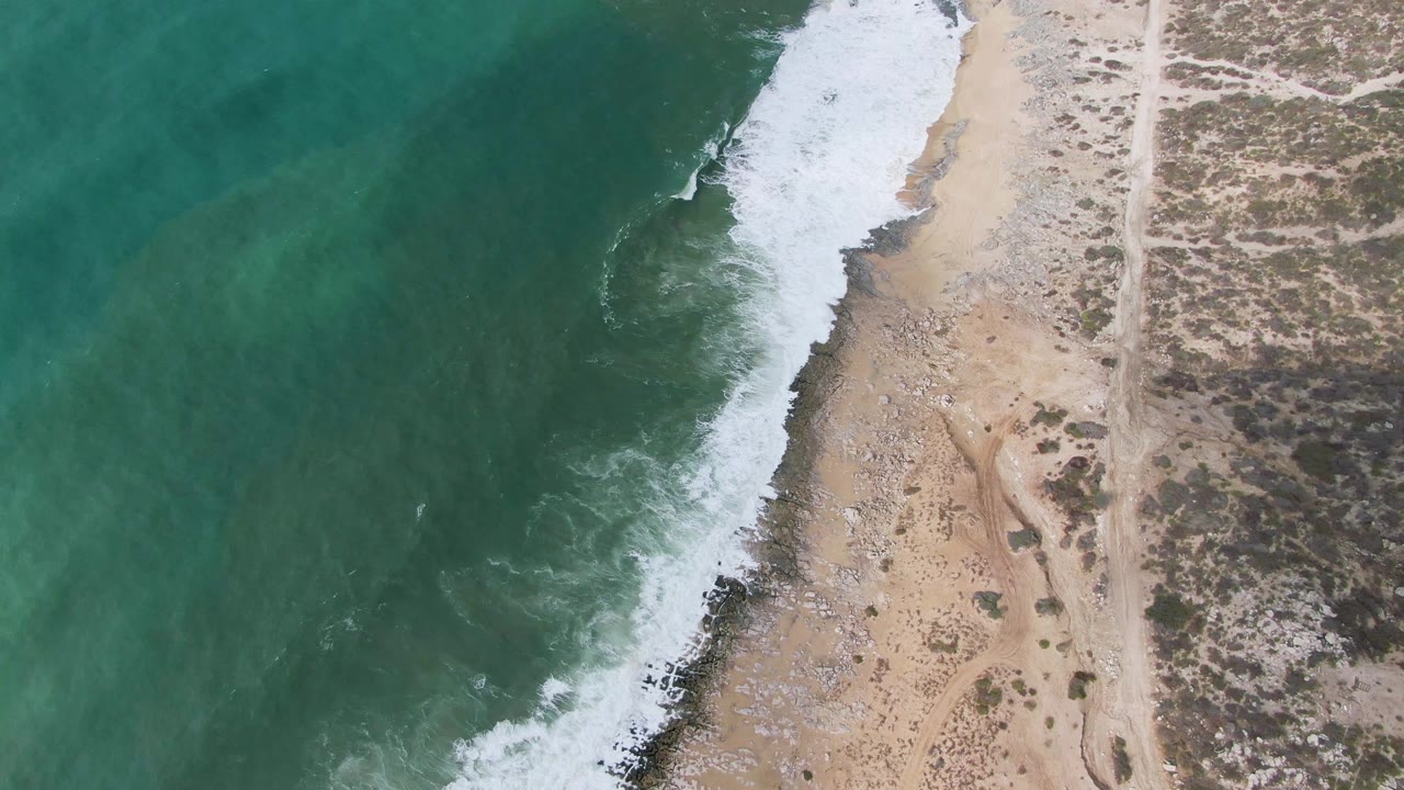 Steep aerial frame from Punta Gorda, Baja California Sur with white surf tracing a diagonal shoreline, pale sand and rocks below it, a coastal track to the right, and no visible people.