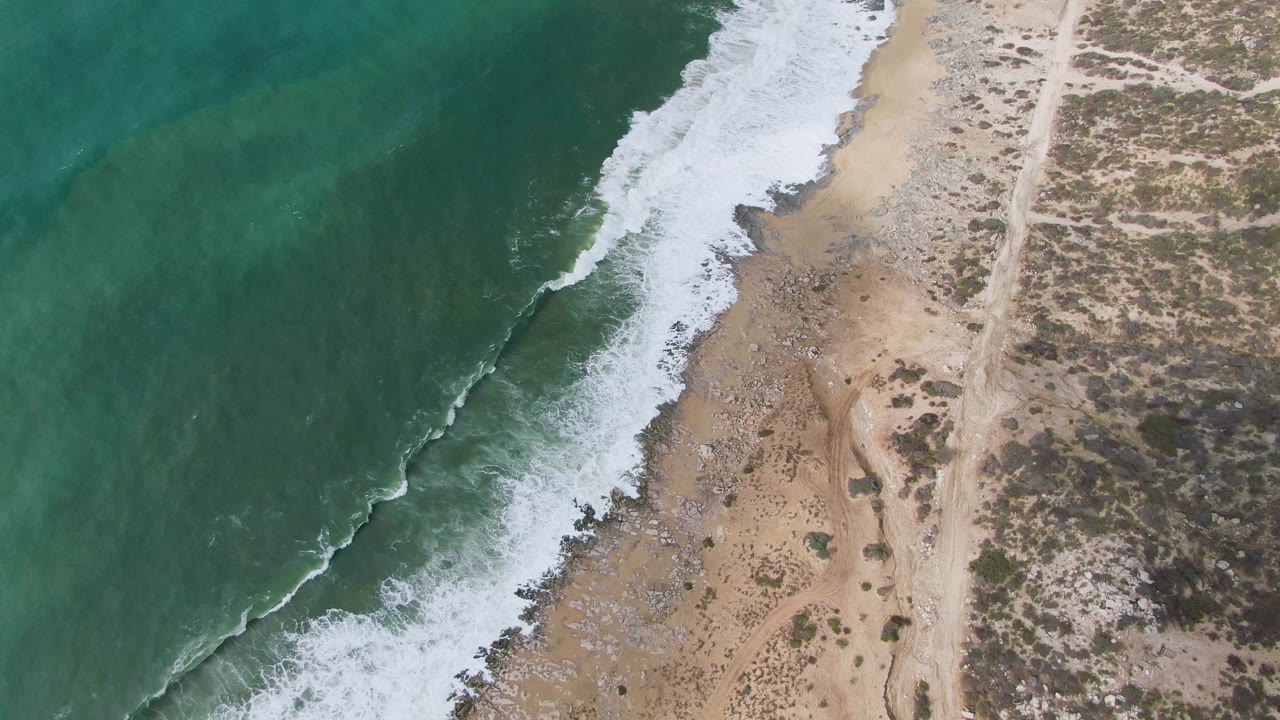Overhead view at Punta Gorda, Baja California Sur, with a white foam band advancing along the sandy beach and a pale dirt track running through sparse vegetation, no visible people.