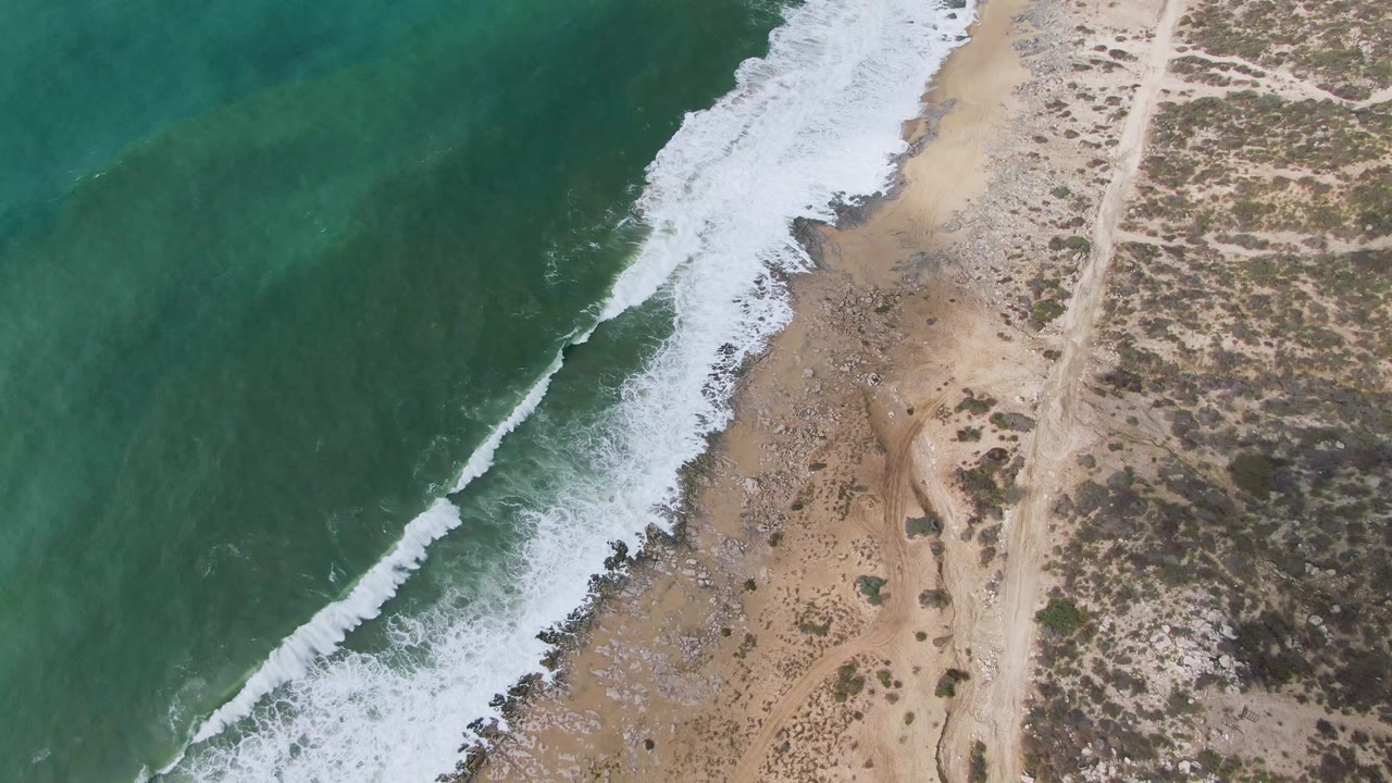 Aerial coastline frame from Punta Gorda, Baja California Sur, showing a darker green swell line offshore, white wash on the beach, sandy ground, and dry coastal vegetation with no visible people.