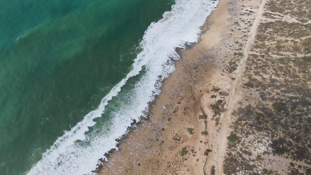 Punta Gorda overhead beach view with foamy surf curling across the lower shoreline, green water to the left, sandy rocky beach in the center, and scrubland to the right with no visible people.