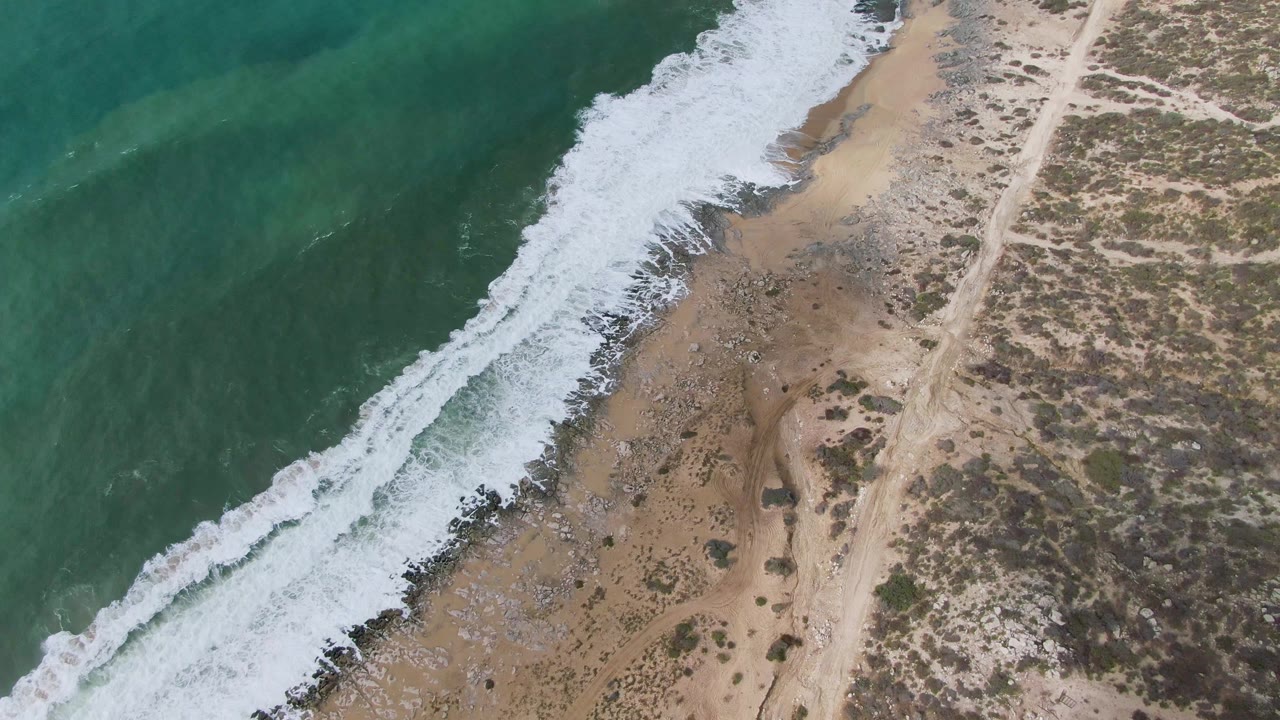Overhead view of green-blue water breaking into white foam along Punta Gorda East Cape sand, with a dirt road through scrubland and no visible people.