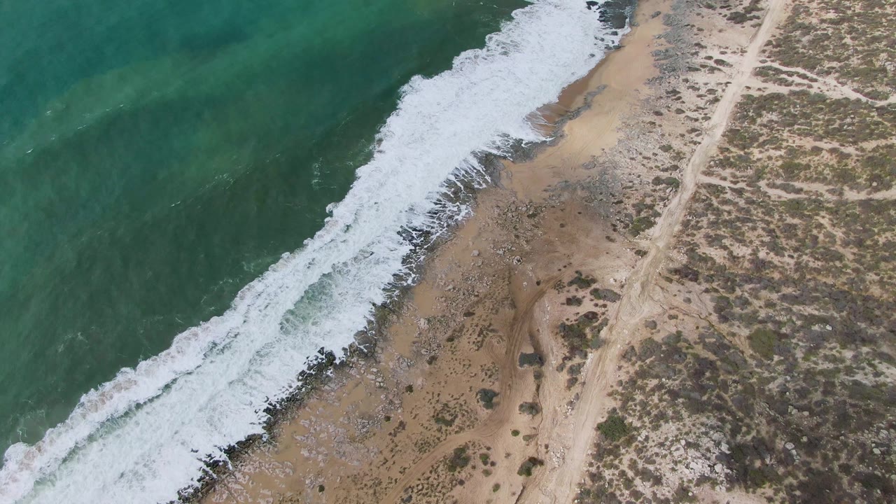 A foamy wave band spreads along the Punta Gorda East Cape shoreline, with tan sand, scattered rocks, a pale dirt road, and no visible people.