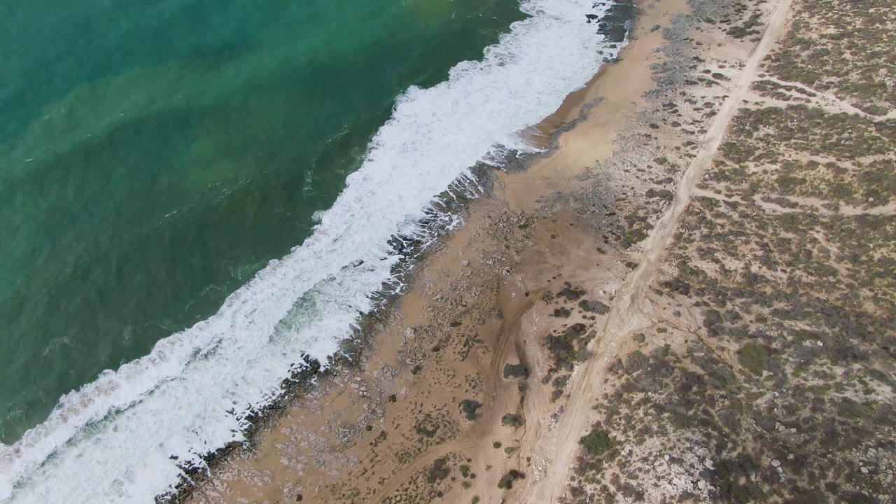 The aerial frame shows Punta Gorda East Cape surf curling diagonally onto sand, with darker rocky patches near the water and no visible people on the beach or road.