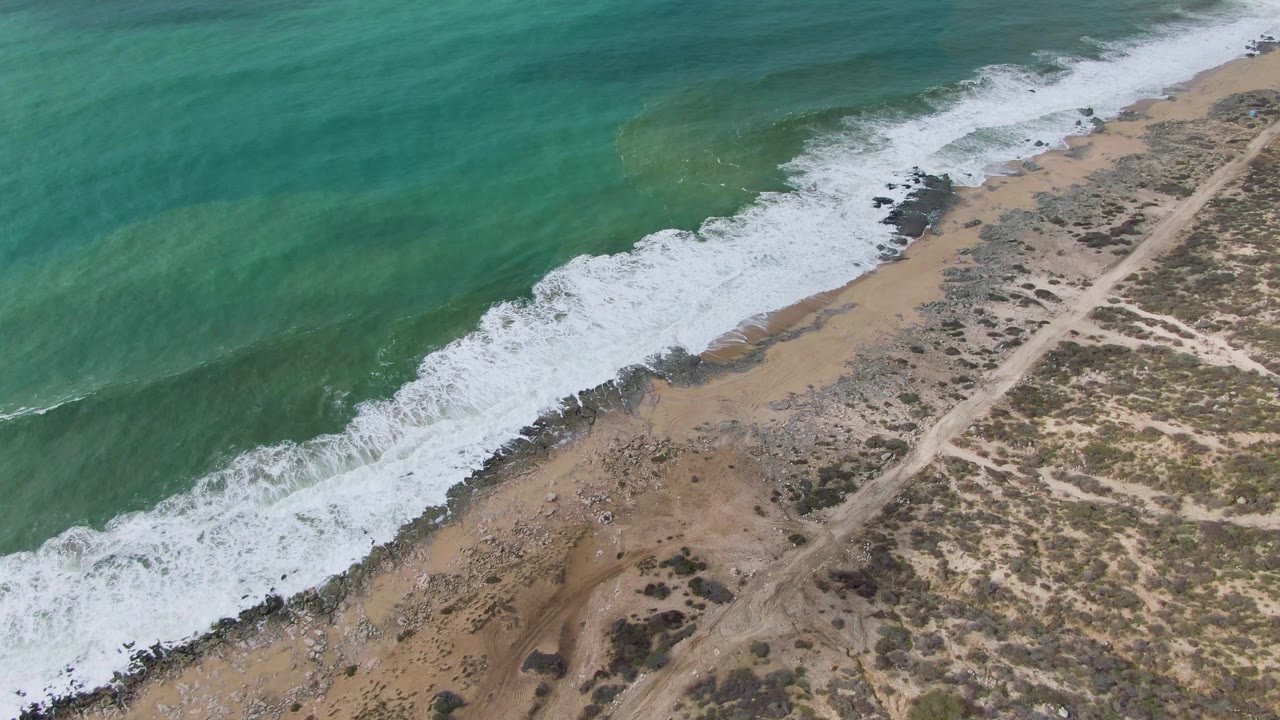 Aerial view of turquoise waves reaching a sandy rocky shore beside a dirt road