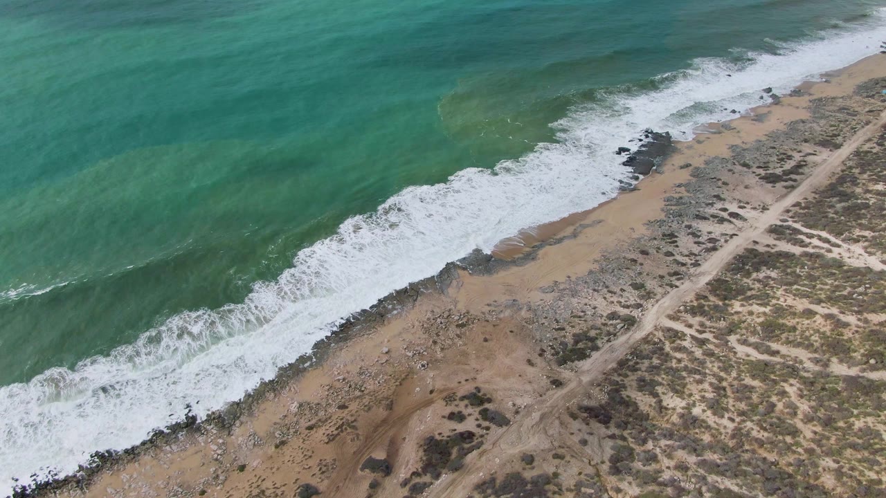 Aerial frame over Punta Gorda East Cape in Baja California Sur, Mexico, with a broad band of white surf advancing across the center of the sandy rocky beach and a pale dirt road running inland, no visible people.