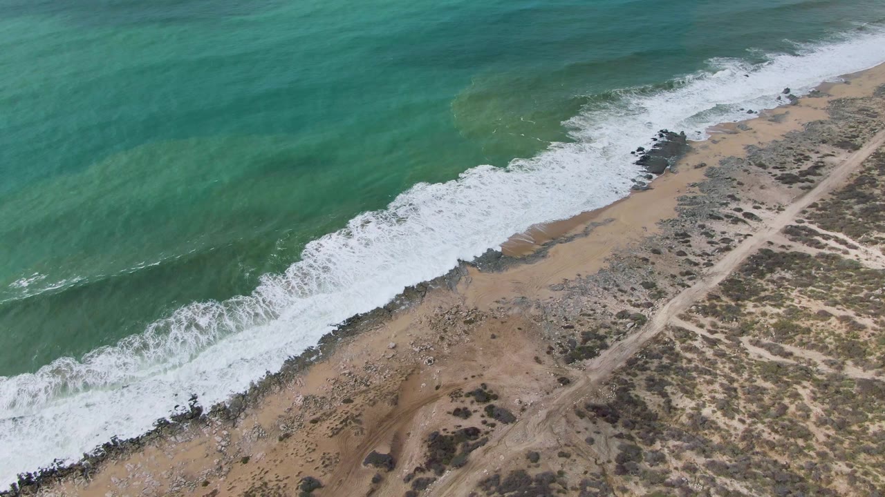 Aerial frame over Punta Gorda East Cape in Baja California Sur, Mexico, where turquoise water meets a diagonal strip of white foam beside dark rocks, sand, scrub, and a dirt road, no visible people.