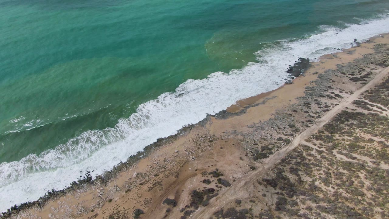 Aerial frame over Punta Gorda East Cape in Baja California Sur, Mexico, showing white water rolling lower across the beach edge, with rocky patches, tan sand, a dirt road, and dry scrub beyond, no visible people.
