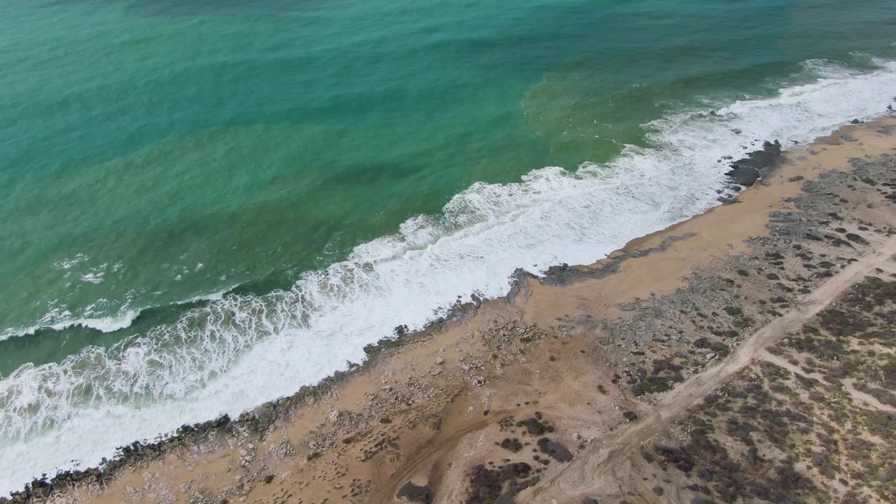 Aerial frame over Punta Gorda East Cape, Baja California Sur, showing turquoise water, a diagonal band of white foam, rocky sand, a dirt path, and no visible people.