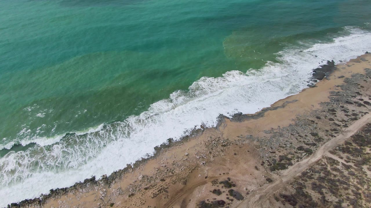 Aerial view of Punta Gorda East Cape surf pushing farther across the tan beach, with dark rock clusters below the foam and no visible people.