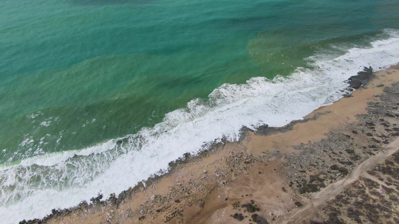 Punta Gorda East Cape shoreline from above, with a brighter white foam band curling along the beach edge, turquoise water offshore, rocky sand inland, and no visible people.