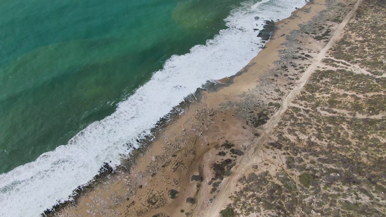 Aerial frame over Punta Gorda, Baja California Sur, showing turquoise water and white surf meeting a sandy, rocky shoreline beside a dirt road, with no visible people.