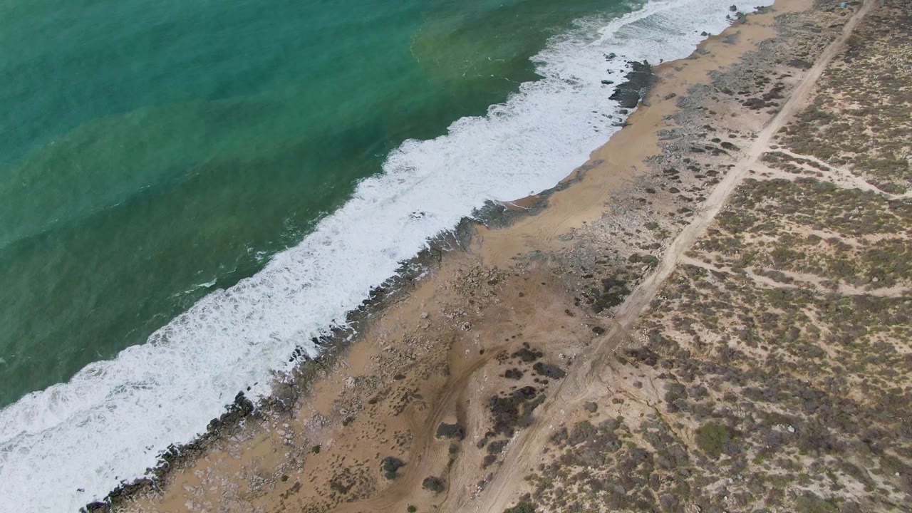 Aerial frame over Punta Gorda, Baja California Sur, with a broader band of white foam pushing onto tan sand and dark rocks near the parallel dirt road, with no visible people.