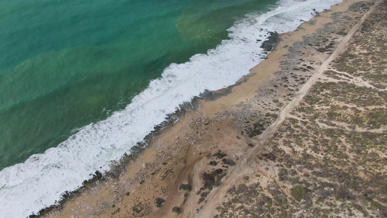 Aerial frame over Punta Gorda, Baja California Sur, showing green-blue water rolling into a bright foam edge along the sandy shoreline and scrubland, with no visible people.