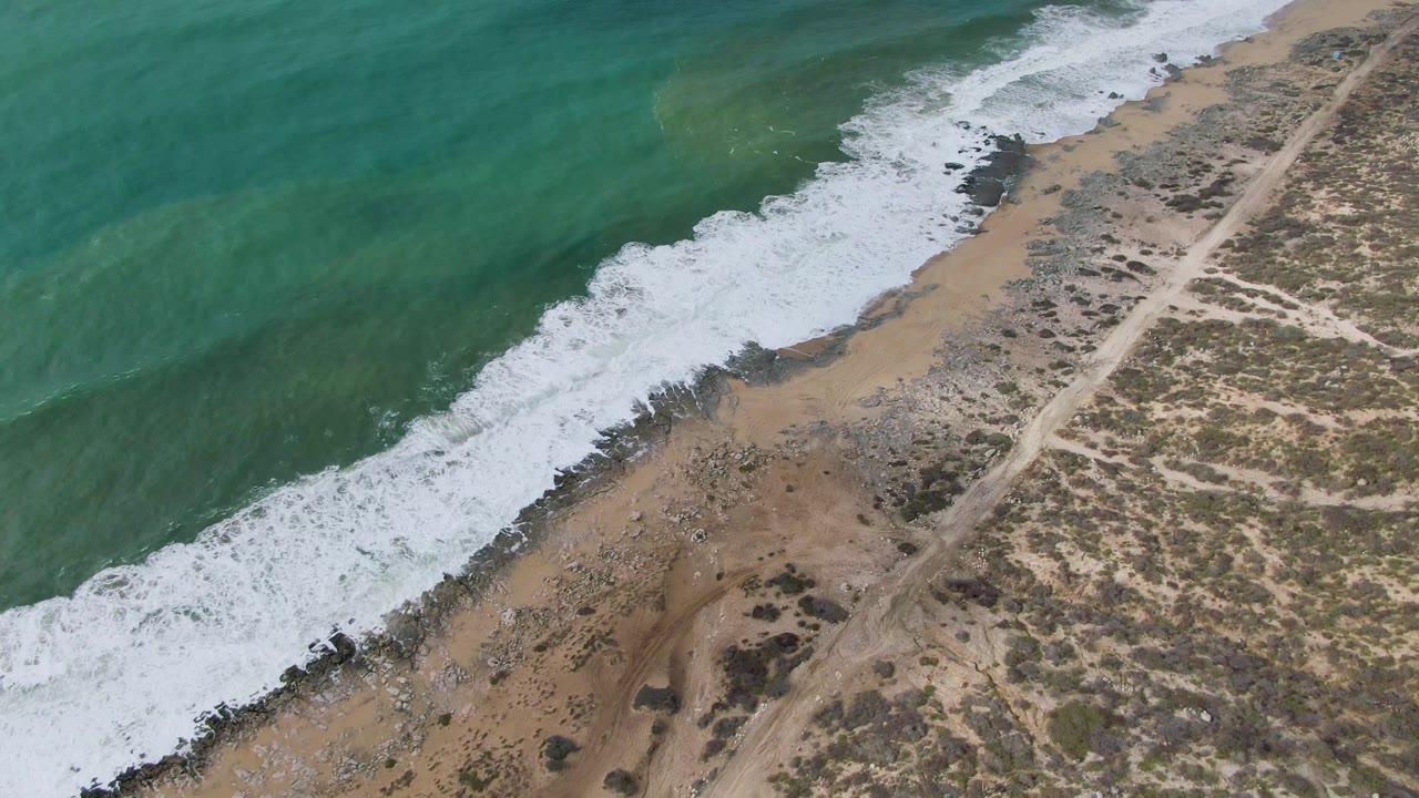 Oblique aerial view of surf washing over rocks along a sandy coastline.